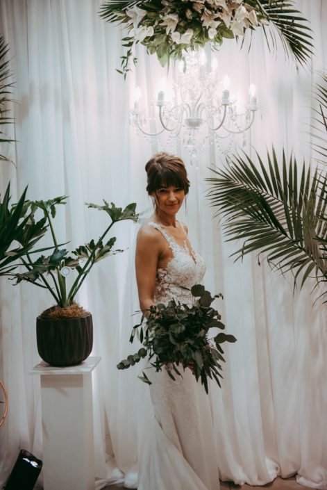 bride with greenery bridal bouquet and tropical plants underneath chandelier and floral arrangement