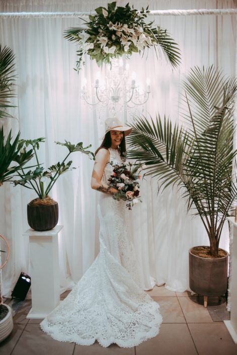 bride with tropical plants in front of white backdrop with chandelier and tropical palm leaf and lily flroal arrangement wearing wedding dress and beige hat and holding a bridal bouquet