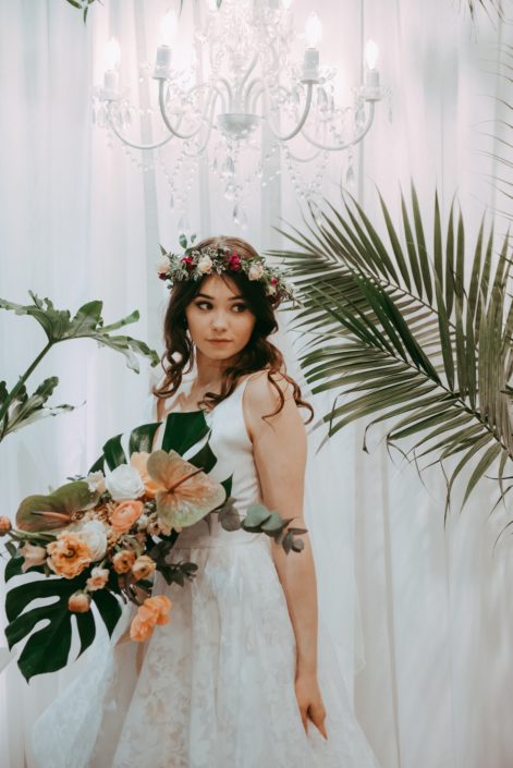 bride with chandelier and white backdrop with tropical plants and bridal bouquet designed with monstera leaves, blush and green anthurium leaves, peach poppies and ranunculus and white roses