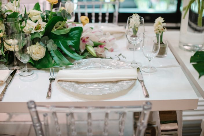 White acrylic table with clear chiavari chairs and white place setting with white floral arrangement and monstera leaves