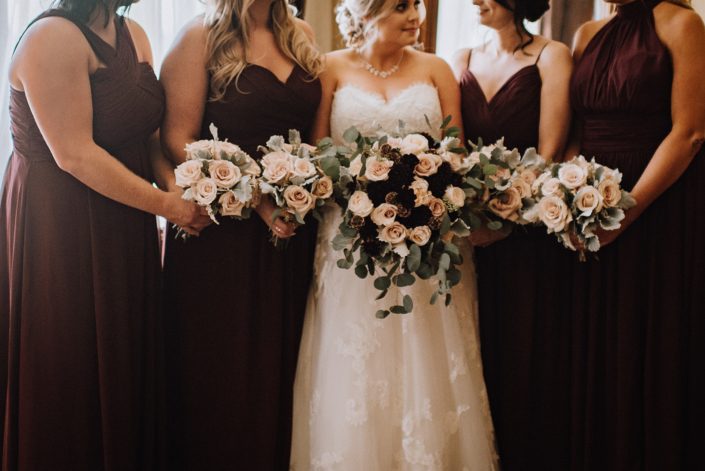 Bride with bridal bouquet of blush roses, burgundy dahlia and eucalytpus and bridesmaids in burgundy with quicksand rose bouquets