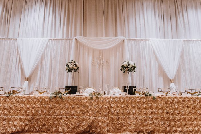 Head table at Cambridge wedding reception with blush rosette table linen and two large and elaborate floral arrangements behind