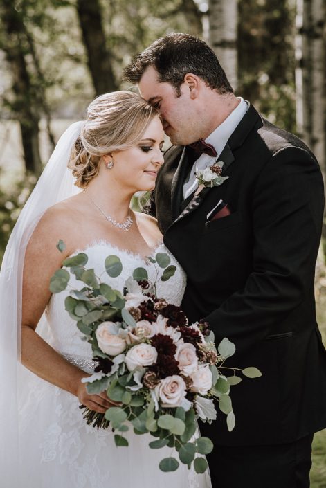 bride in white wedding dress and long veil holding bridal bouquet of blush roses and burgundy dahlia and eucalyptus with groom in black suit with burgundy bow tie and white rose boutonierre
