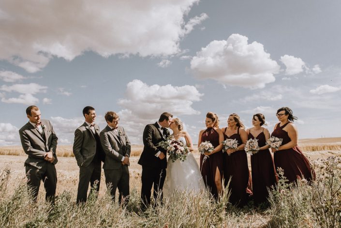 Wedding party in summer wheat feild in grey suits and burgundy bridesmaid dresses with bouquests designed with blush roses and burgundy dahlia