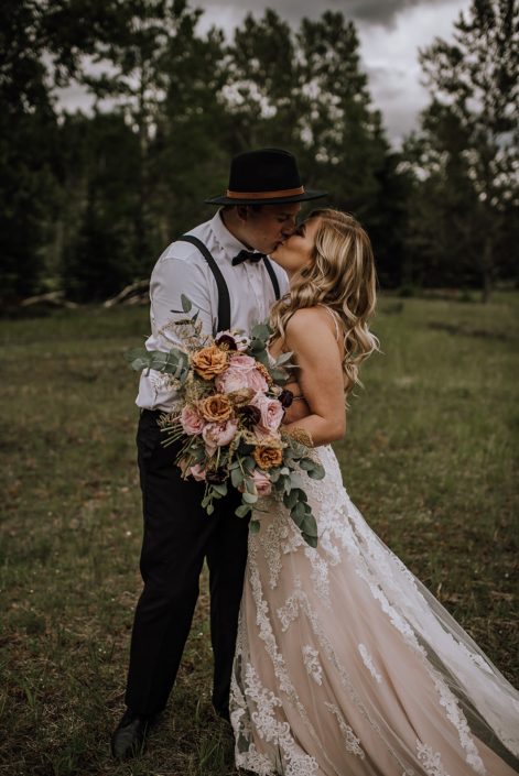 bride and groom in black hat kissing holding a bridal bouquet in an organic shae with toffee roses, pik oahra garden roses and cinerea eucalytpus