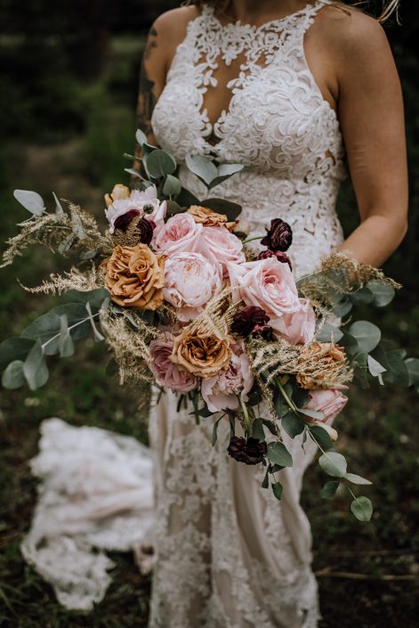 bride in white lace wedding dress holding organic oblong bridal bouquet designed with gold plumosa, cinerea eucalyptus, toffee roses, plum ranunculus, pale pink peony and pink ohara garden roses
