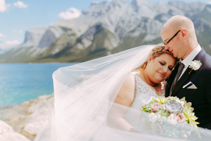Bride and groom with veil in front of a lake in the summer with a succulent boutonniere and bridal bouquet