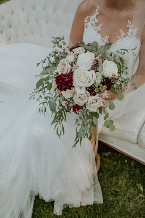 close up photo of bridal bouquet abd wedding dress on an antique ivory sofa designed with burgundy dahlia, ivory and blush roses and eucalyptus