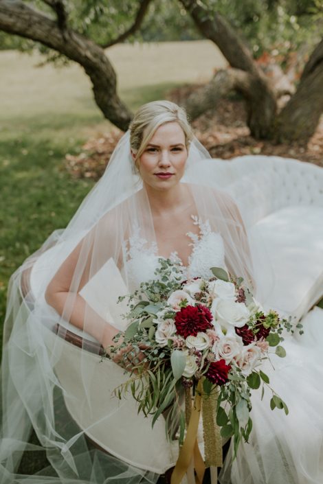 bride sitting on an antique ivory sofa holding a bridal bouquet designed with ivory, blush and burgundy and trailing gold sequin ribbons wearing a cathedral length veil