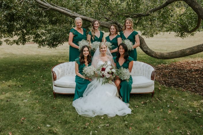 bride and bridesmaids in forest green sitting on an antique ivory and wood sofa holding bridesmaid bouquets of babies breath and brides bouquet of burgundy dahlia, blush roses and ivory roses and eucalyptus