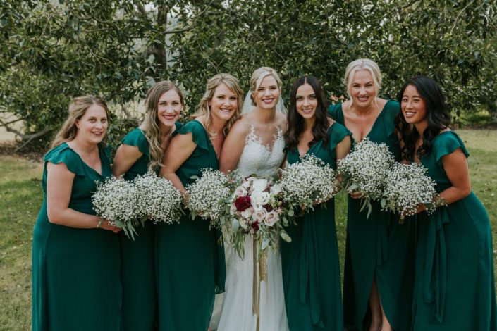 bride and bridesmaids in forest green dresses holding bridesmaids bouquets of babies breath with eucalyptus and bridal bouquet of burgundy dahlia, blush and white roses and eucalyptus greeenery with trailing silk and sequin gold ribbons