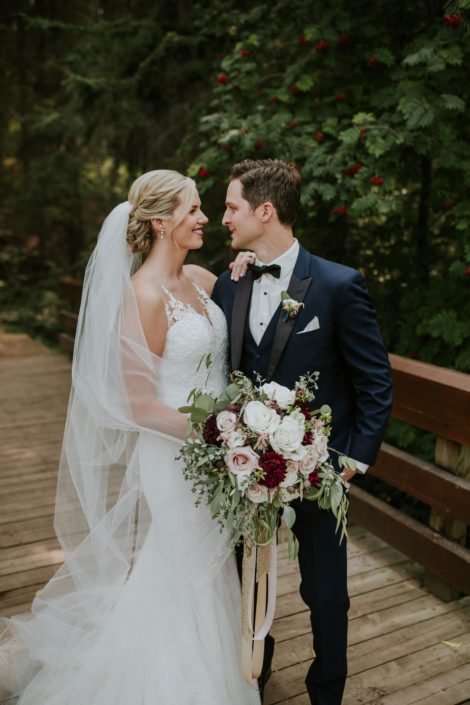 bride wearing a white wedding dress and cathedral veil with groom in navy tuxedo holding a bridal bouquet made with burgundy dahlia, white roses and blush roses and eucalyptus greenery