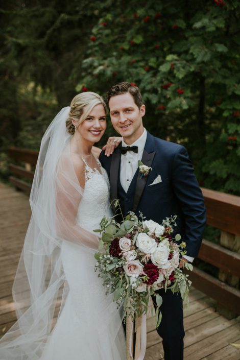 bride and groom on a bridge in the forest for s summer wedding holding a bridal bouquet designed with burgundy dahlia and blush and white roses and eucalyptus greenery
