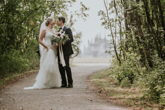 bride and groom on path in the summer with a wedding dress and veil holding a bridal bouquet of eucalyptus and ivory spray roses and burgundy dahia