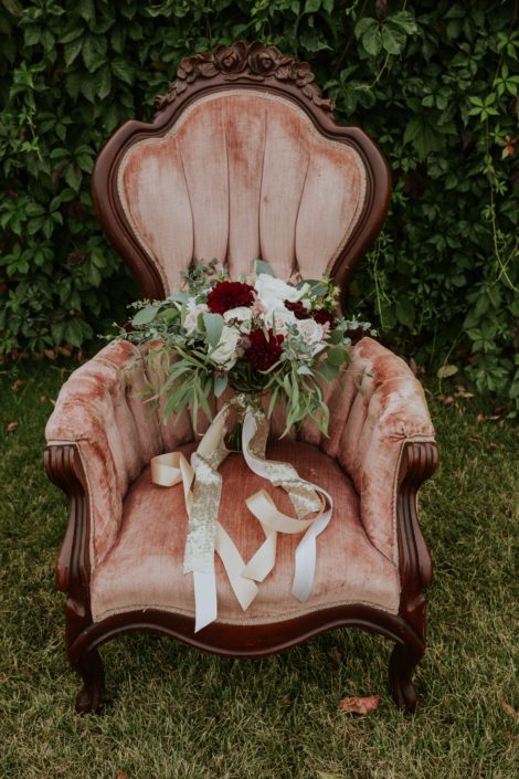 antique dusty rose chair with bridal bouquet that has trailing gild sequin and blush silk ribbons designed with burgundy dahlia, blush and ivory roses