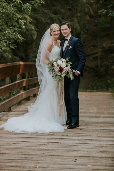 bride and groom on a bridge with wedding dress and veil and navy suit holding a bouquet designed with eucalyptus, burgundy dahlia and white and blush roses