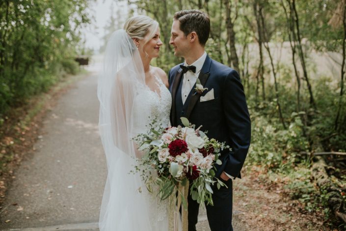 Bride with cathedral length veil and lace wedding dress with groom in navy tux with spray rose boutonniere holding a burgundy, blush and ivory bridal bouquet with eucalyptus collar