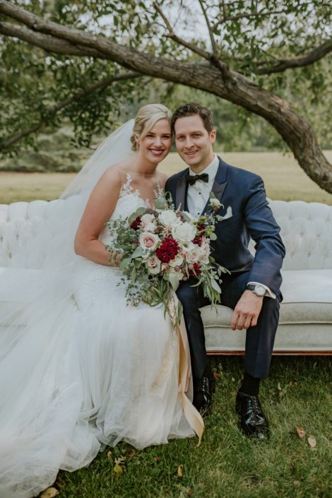 bride with veil and wedding dress and groom in navy tux sitting on an antique ivory sofa holding a blush, burgundy and ivory bridal bouquet with fresh eucalyptus greenery collar