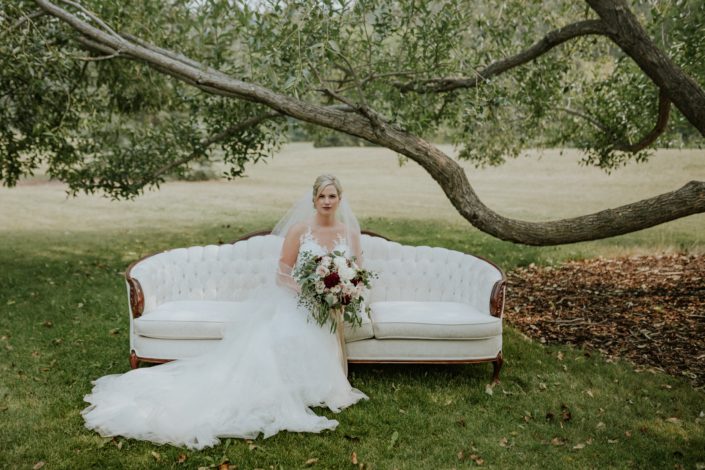 bride sitting in a park under an old tree on an ivory sofa holding a bridal bouquet in blush, white and burgundy with eucalyptus greenery