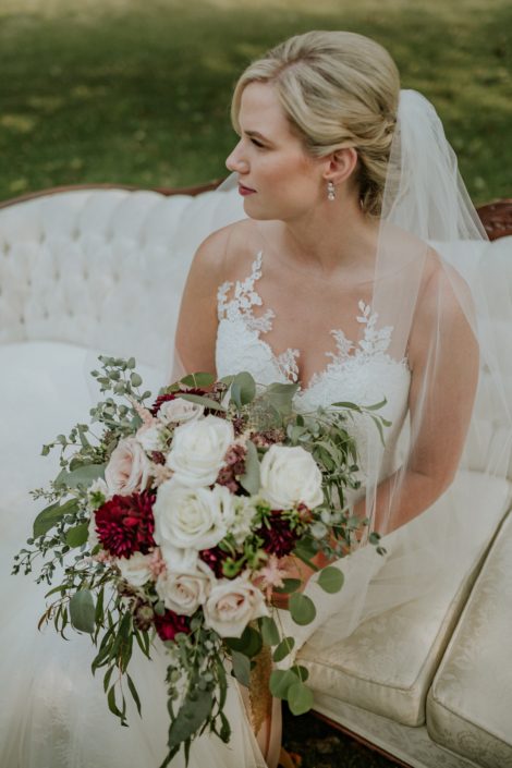 bride with a bridal bouquet designed with eucalyptus and blush and ivory roses and burgundy dahlia wearing pearl earings and an ivory veil