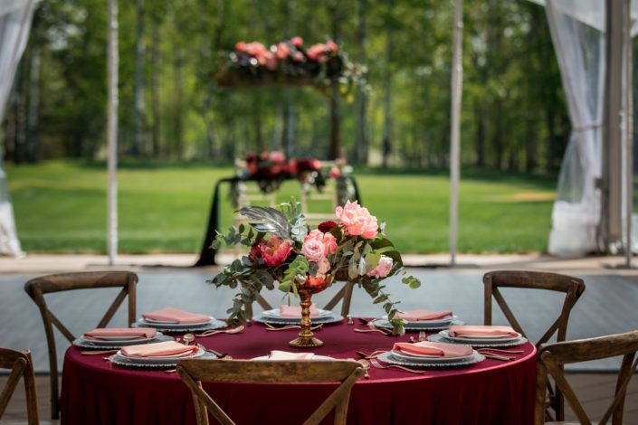 Gold compote centerpiece with black monstera leaves, coral charm peony, burgundy dahlia and cinerea eucalyptus ona burgundy tablecloth with vineyard chairs