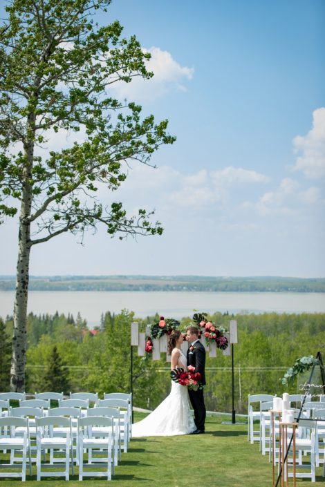 wedding ceremony set up at hilltop wedding venue in sylvan lake with archway backdrop of coral and burgundy and black with bride and groom and bridal bouquet