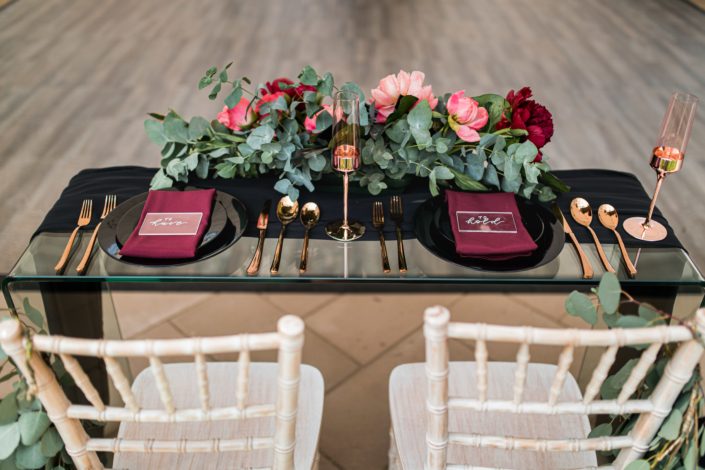 clear glass sweetheart table with limewash chiavari chairs, black dinnerware and gold wine glasses and cutlery with a floral arrangement of penies in coral and burgundy and cinerea eucalyptus