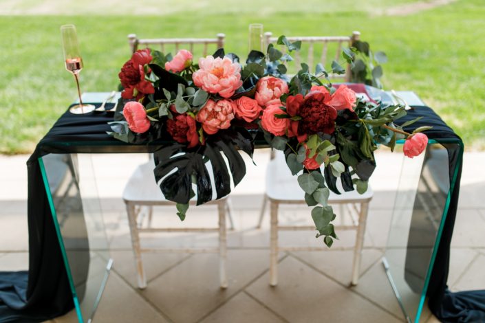 clear glass sweetheart head table with black voile runner and gold wedding glasses with floral arrangement designed with black monstera, burgundy and coral peony, coral amsterdam roses and cinerea eucalyptus
