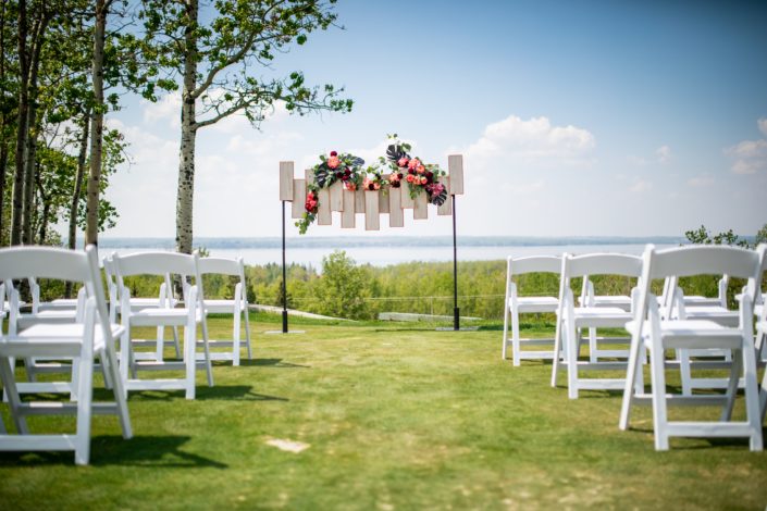 archway florals with black monstera coral peony and burgundy peony with geometric archway backdrop and white folding chairs overlooking sylvan lake