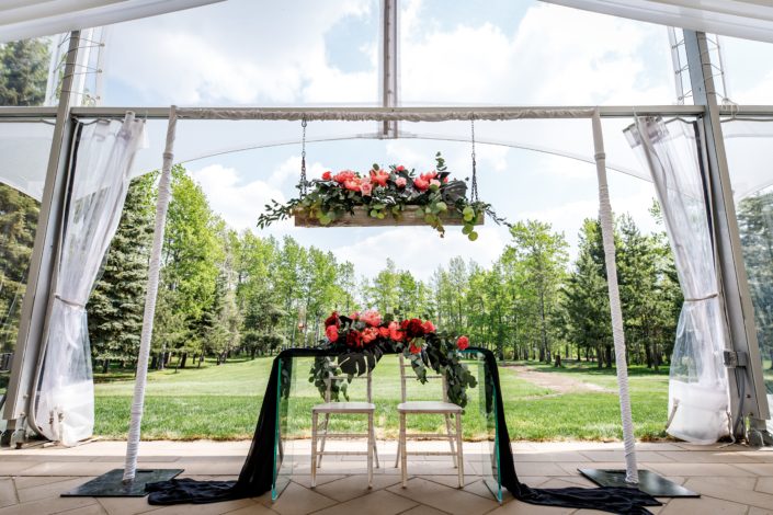 clear wedding tent at Hilltop Wedding center sweetheart table of clear glass with black voile runner and fresh floral arrangements designed with black monstera leaves, coral charm peonies and burgundy peonies
