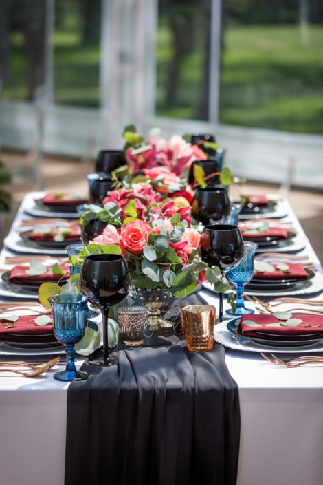 rectangular table with black table runner and wine glasses and three compote style centerpieces in coral and burgundy