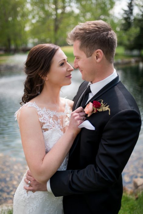 bride in white lace wedding dress with groom in black tuxedo wearing a boutonierre with peach ranunculus and burgundy rose and gold plumosa accent