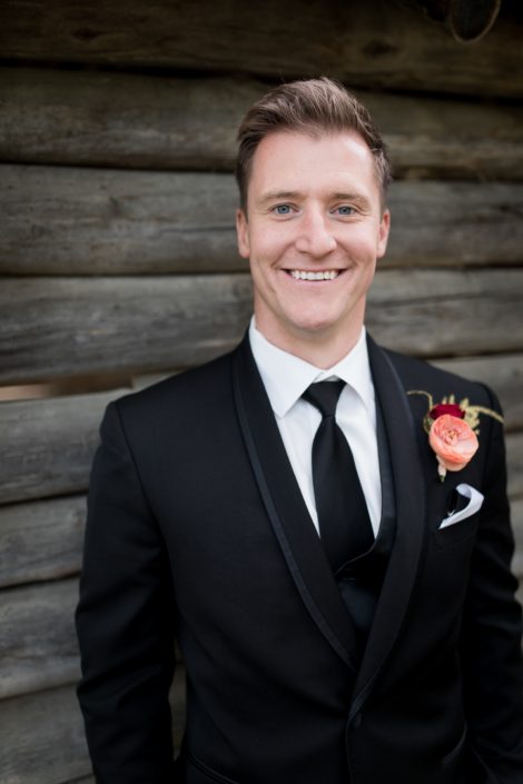 Groom in black tux with salmon ranunculus and burgundy spray rose boutonniere