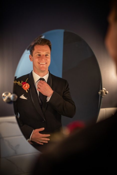 groom in grooms room at hilltop wedding center in black tux with coral and burgundy boutonierre