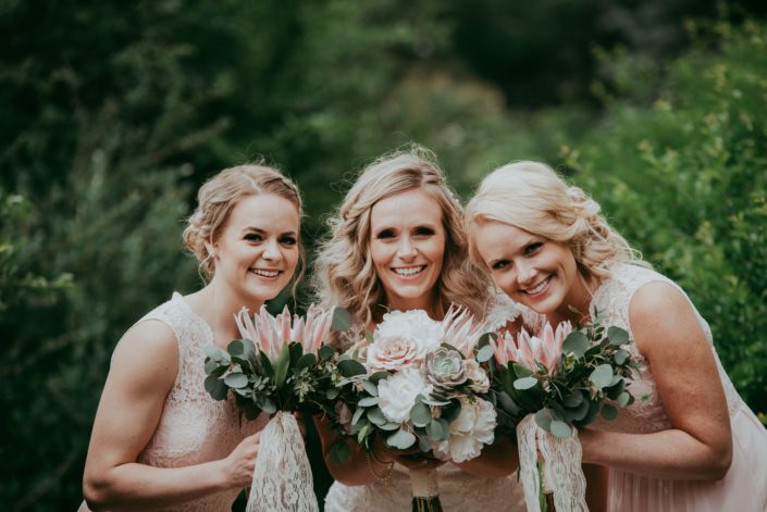 Blond bride and bridesmaids in blush wedding dresses holding bouquets of silver dollar eucalyptus and pale pink king protea with white peony and succulents