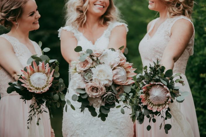 bride and bridesmaids in blush lace holding bouquets of king protea, eucalyptus and white peony and blush roses