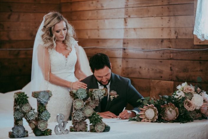 bride and groom at signing table at wedding with succulent monogram N and K and bouquets of King protea and eucalytpus