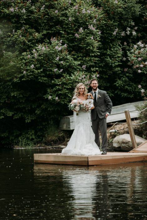 bride and groom on dock at pond holding spaniel puppy dog with bridal bouquet in white and blush and eucalyptus