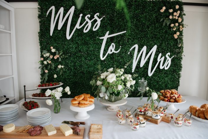 Boxwood backdrop for miss to mrs bridal shower overlooking dessert table with donuts, muffins ad charcuterie board and floral arrangements in blush, white and greenery