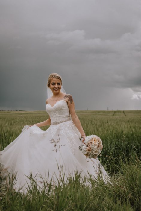 bride in a summer wheat field in alberta holding a bridal bouquet of blush roses and white hydrangea and dusty miller
