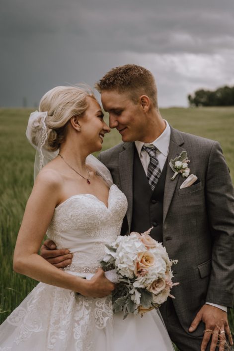 bride and groom in stormy alberta weather in wheat field holding bridal bouquet of white hydrangea and blush rose and dusty miller bouquet