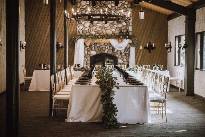 Wedding reception decor with white linens and black ine glasses and charger plates decorated with fresh eucalyptus garland and gold chiavari chairs