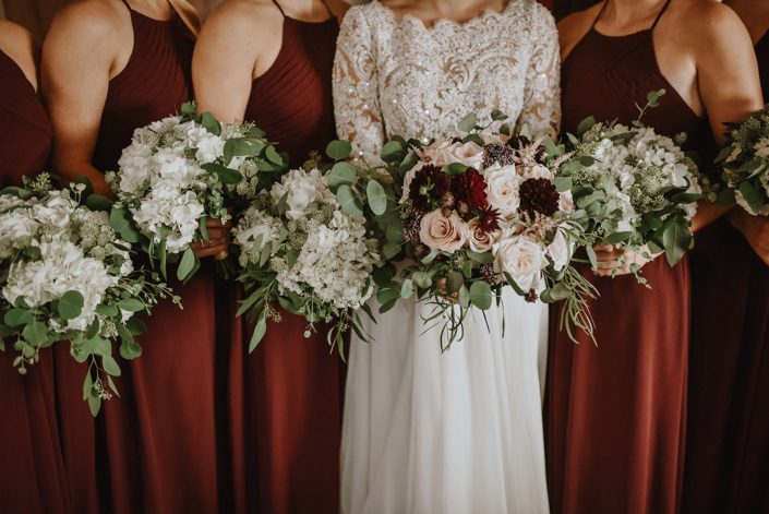 bride with bridal bouquet of blush and burgundy dahlia and roses and bridesmaids in burgundy holding bouquets of white hydrangea and astrantia and eucalyptus