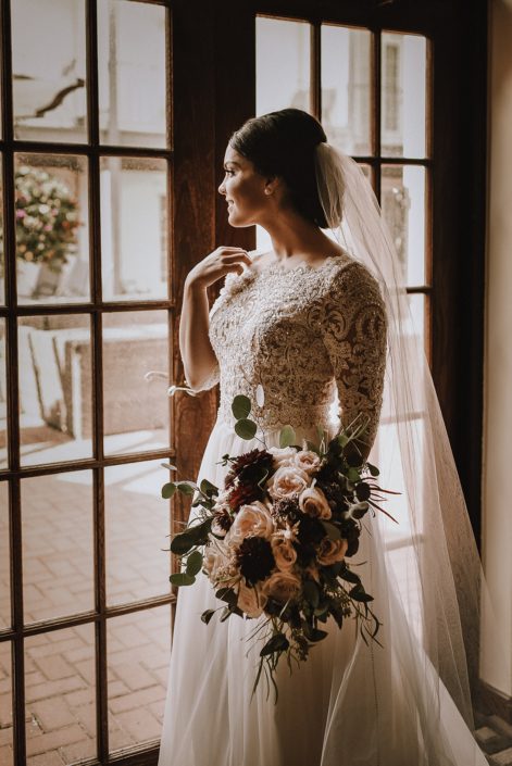 bride in long sleeved lace top wedding dress wth cathedral veil holding a bridal bouquet designed with burgundy dahlia, ivory garden roses and blush garden roses