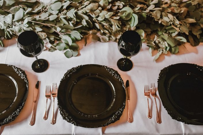 Wedding table setting with a white linen, black wine glasses and charger plates, gold cutlery and eucalyptus garland runner