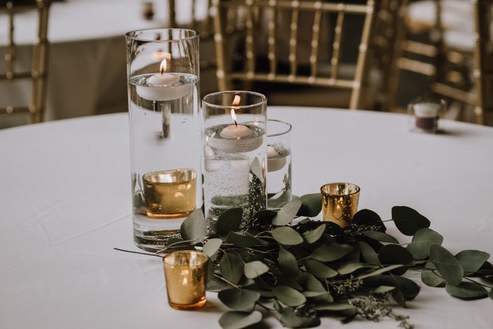 White linens on table with clear cylinder vases and candles and fresh eucalyptus at base