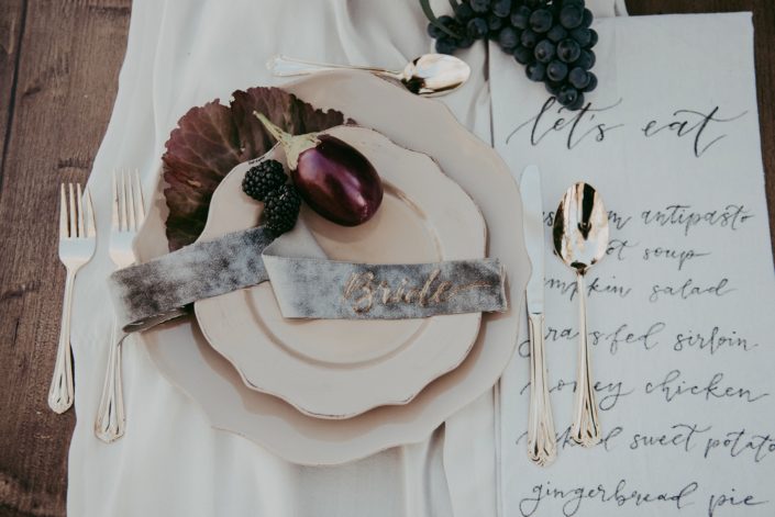 Place setting at harvest autumn table with grapes and aubergine and velvet placecards for bride with gold cutlery and beige dinnerware on blush silk tablerunner