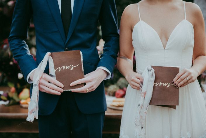 Bride in truvelle wedding dress and groom in navy suit holding leather and silk ribbon bound vow books