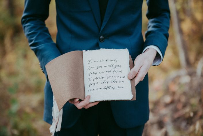 Groom in navy suit holding modern calligraphy leather bound vow book