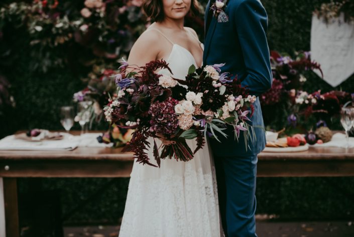Close up of bridal bouquet designed with burgundy amarathus and hydrangea, blush spray roses and carnations and purple clematis and groom in navy blue suit in front of a boxwood backdrop